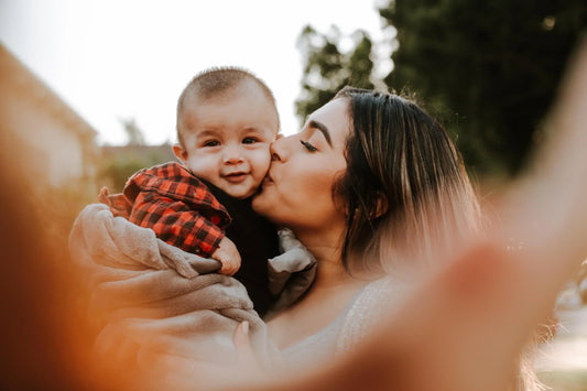 A mother kisses a baby boy's cheek. The baby is wearing a red and black plaid shirt and smiling. They are outdoors with blurred greenery and a building in the background, captured in warm sunlight.