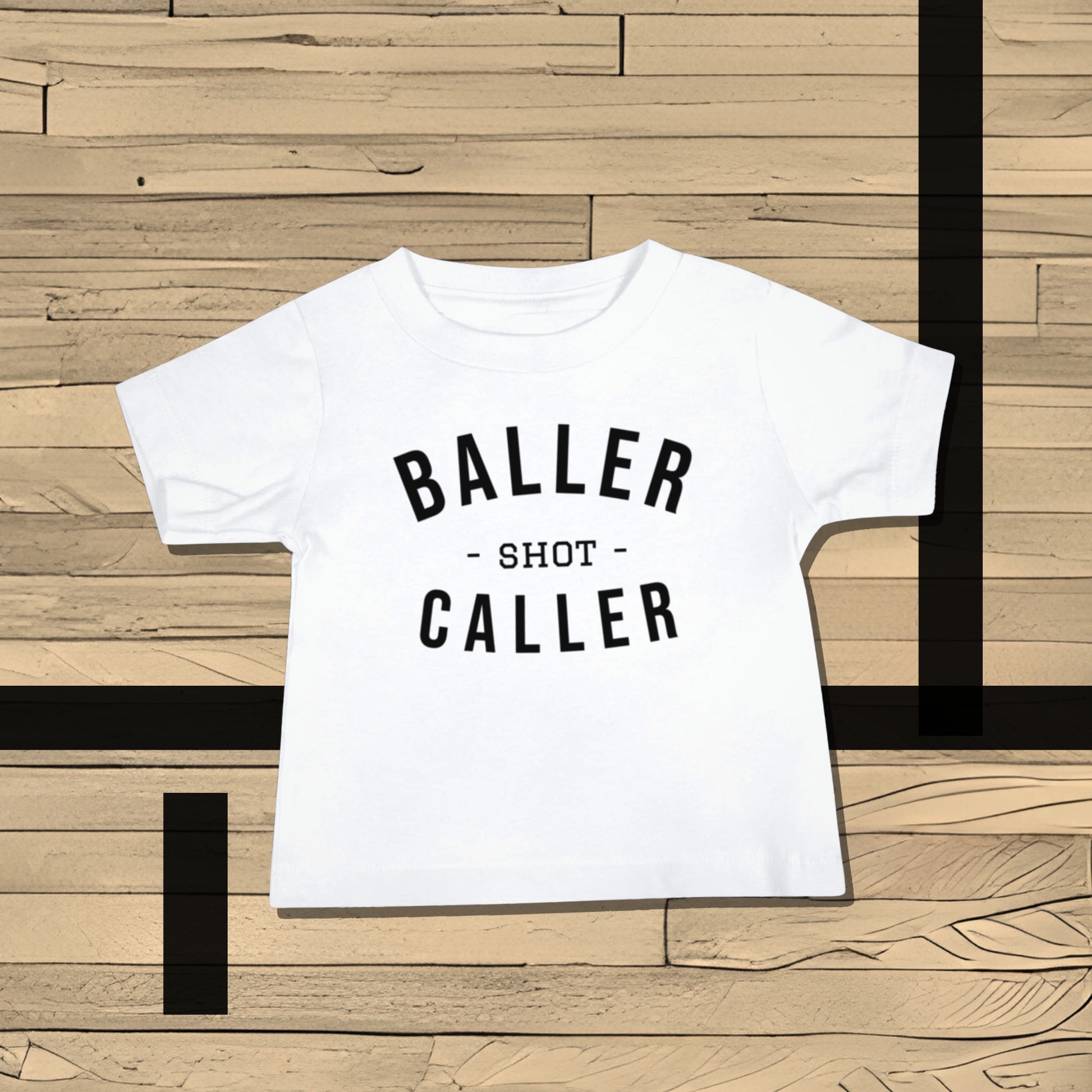 White baby t-shirt with "BALLER - SHOT - CALLER" text on a wooden basketball court. A bold attitude baby clothes t-shirt design for little shot callers, making it a unique baby shower gift from Born Brash for new parents who love sports.