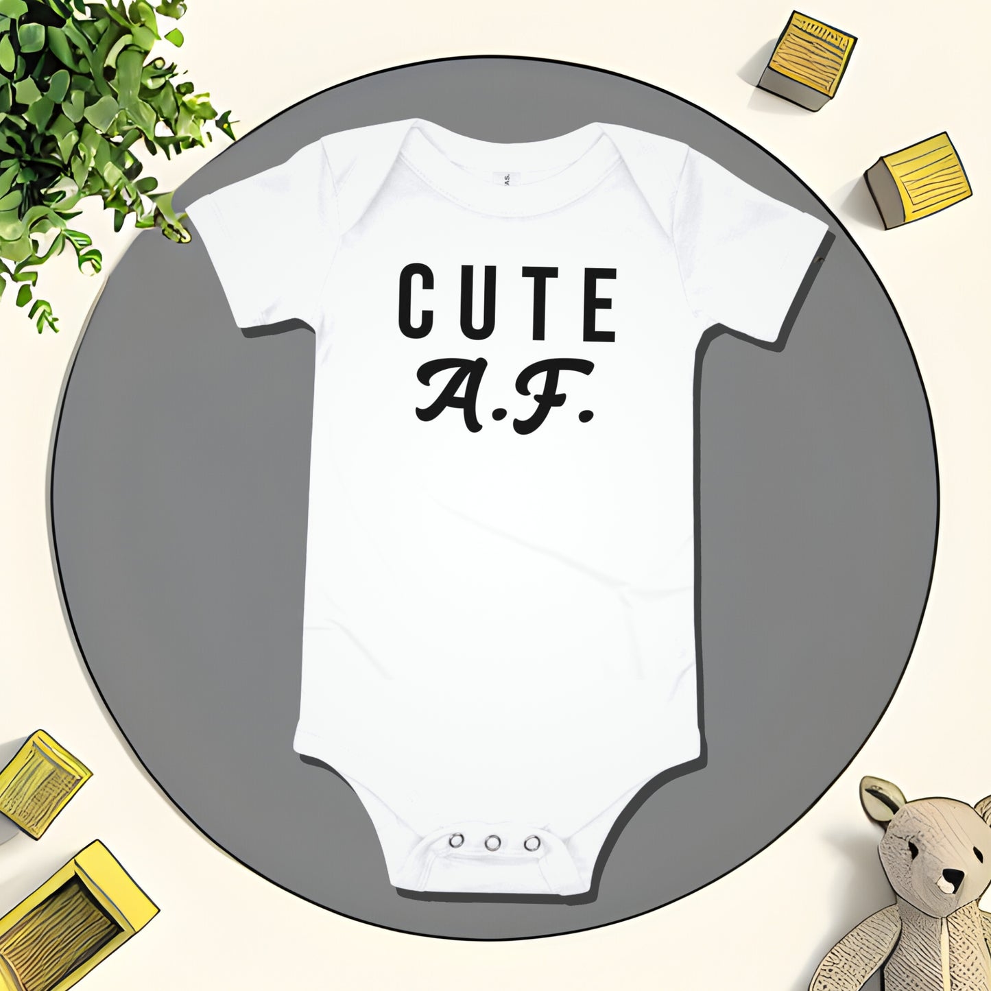 White baby bodysuit with "CUTE A.F." text on a gray circular mat with toy and plant illustrations. A bold attitude baby clothes design with a funny unapologetic statement from Born Brash.