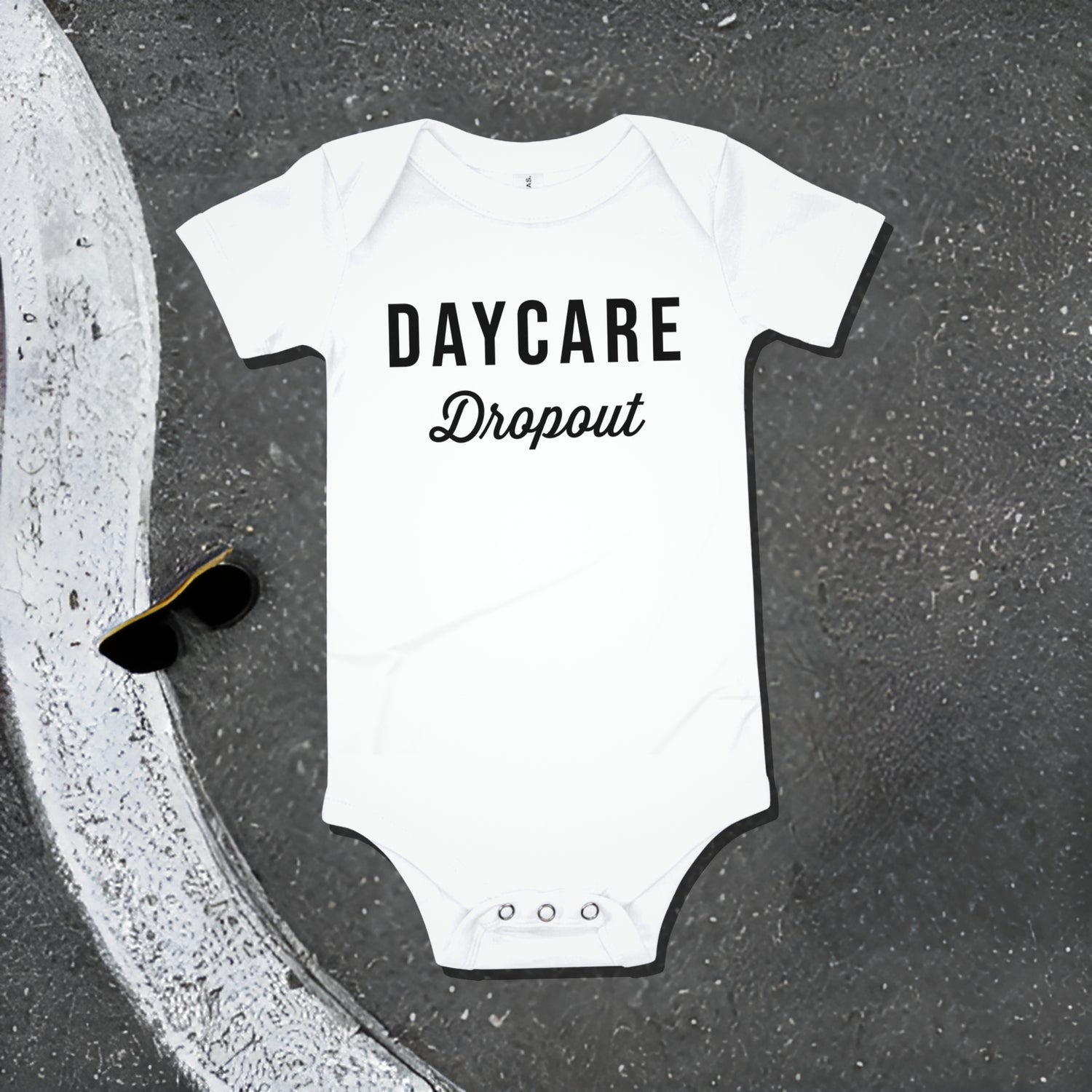 White baby bodysuit with "DAYCARE Dropout" text on an asphalt background with road line and sunglasses. A funny humorous and rebellious baby clothes design about daycare from Born Brash.