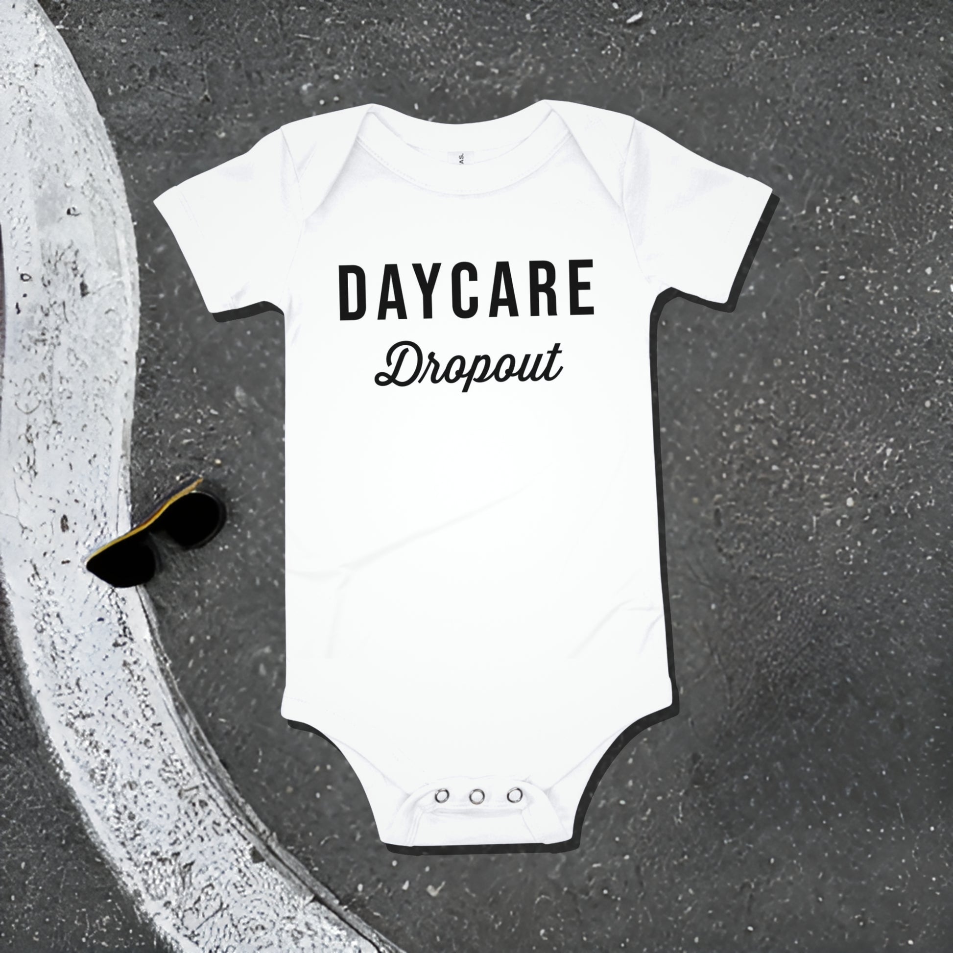 White baby bodysuit with "DAYCARE Dropout" text on an asphalt background with road line and sunglasses. A funny humorous and rebellious baby clothes design about daycare from Born Brash.