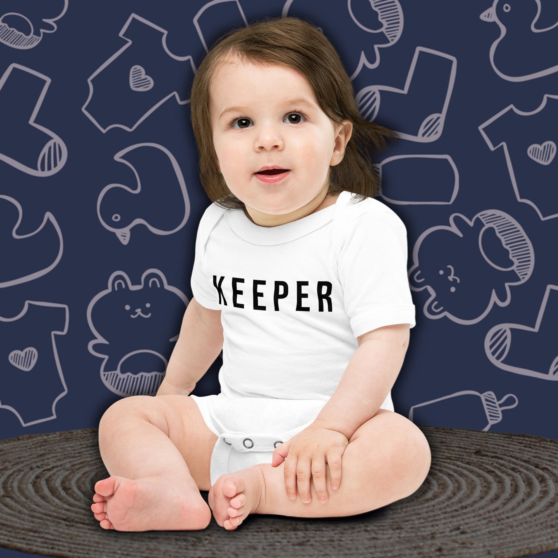 A cute baby with dark hair, sitting and looking forward, wearing a cute, white Born Brash bodysuit that says 'Keeper'. The background is dark blue with white doodle patterns of baby bodysuits, baby toys, and baby bottles. Perfect for a cute newborn gift or pregnancy announcement.