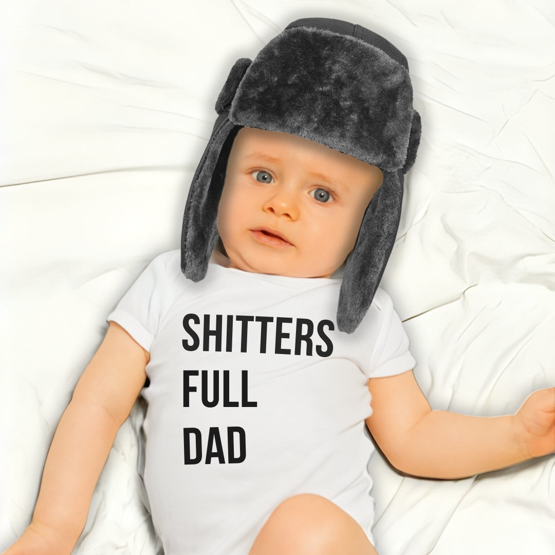 Baby wearing a white "SHITTERS FULL DAD" bodysuit and black fuzzy hat. A funny humorous baby outfit perfect for memorable baby photos or as a hilarious baby shower gift for new dads from Born Brash.