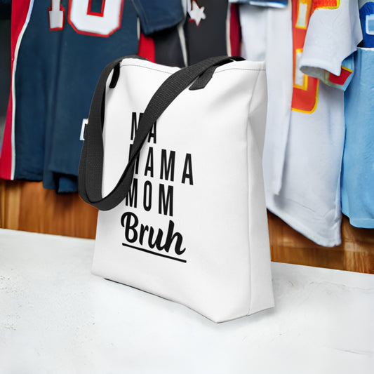 White tote bag with "MA MAMA MOM Bruh" text on a surface with blurred sports jerseys. A funny humorous parenting diaper bag, making it a great baby shower gift for expecting or new moms from Born Brash.