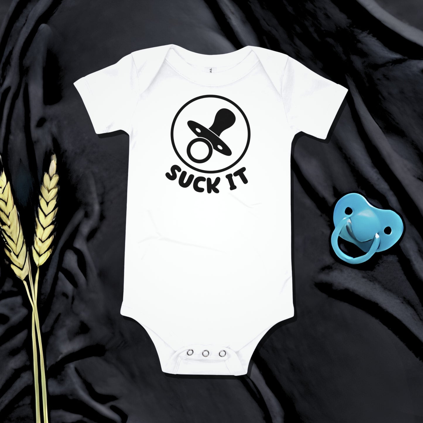 White baby bodysuit with a pacifier graphic and "SUCK IT" text on a dark background with a pacifier and wheat. A funny humorous and irreverent baby clothes design from Born Brash.