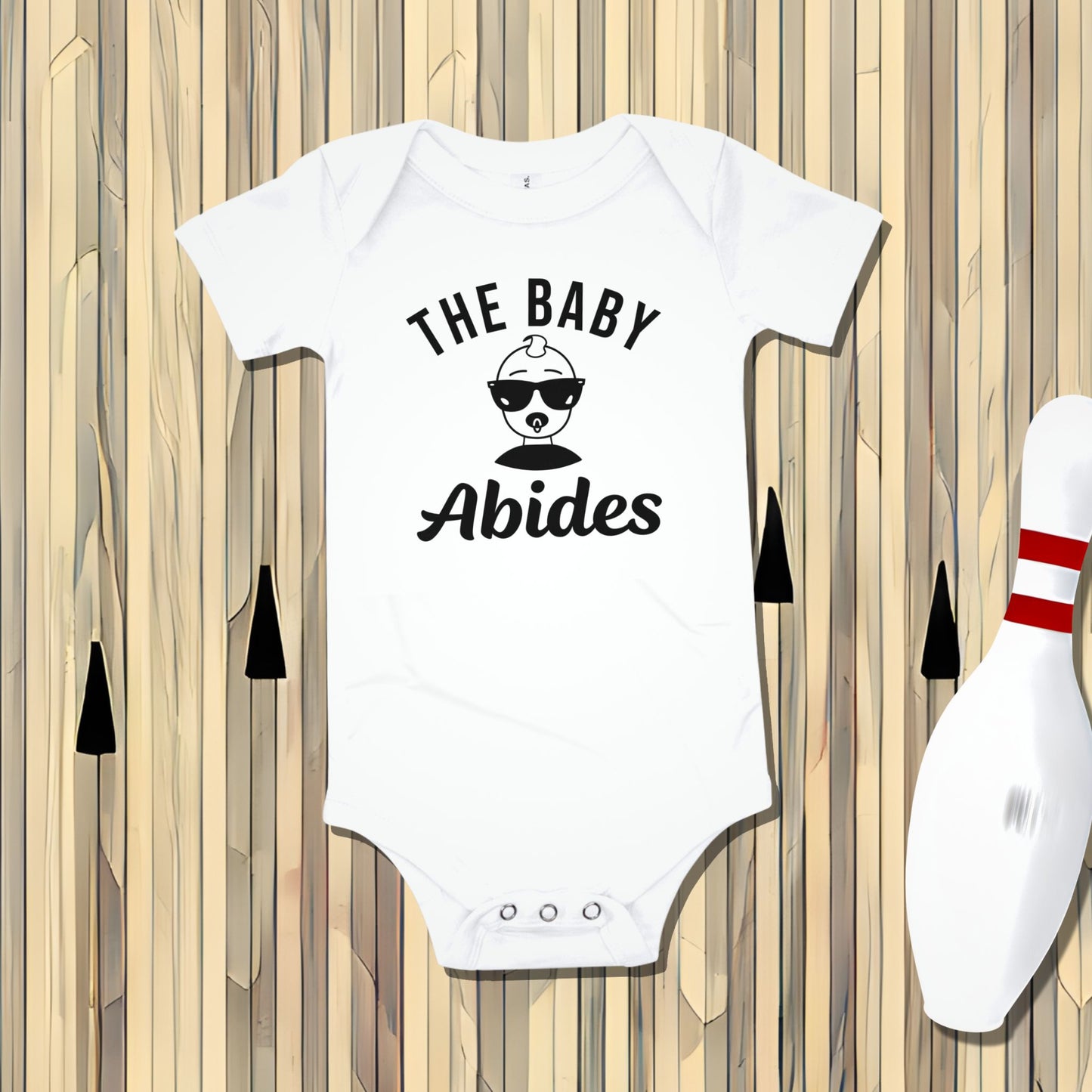 White Born Brash baby bodysuit that says 'THE BABY Abides' with a cool baby wearing sunglasses. The bodysuit is laid flat on a bowling alley lane background with a bowling pin. Funny baby clothes, The Big Lebowski inspired outfit, unique baby gift for fans.