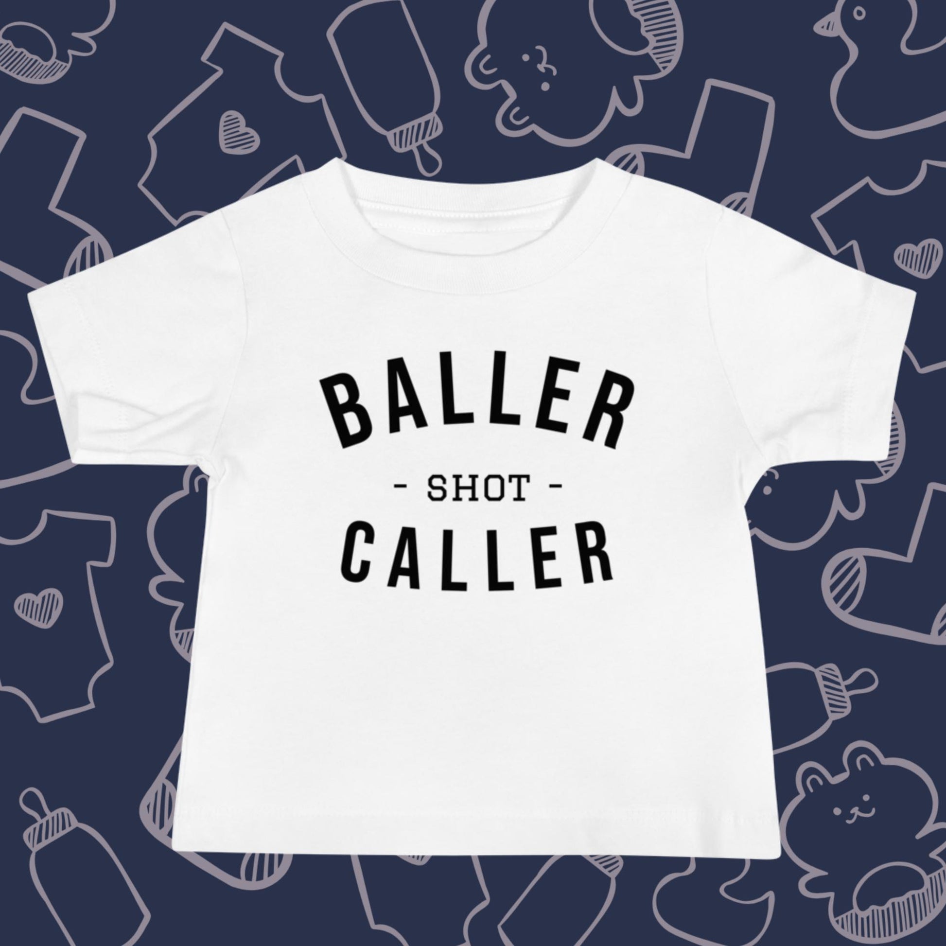 Funny, white baby t-shirt featuring the playful text "BALLER SHOT CALLER" against a whimsical, blue backdrop of ducks, socks, stuffed animals, baby bodysuits, and bottles