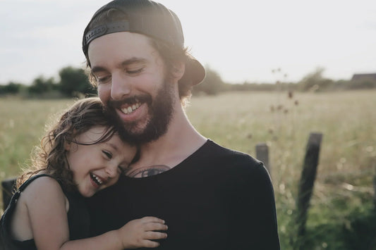 A father holds his young daughter outdoors, both smiling and laughing in warm, blurred lighting in a grassy field.