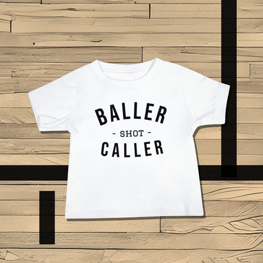 White baby t-shirt with "BALLER - SHOT - CALLER" text on a wooden basketball court. A bold attitude baby clothes t-shirt design for little shot callers, making it a unique baby shower gift from Born Brash for new parents who love sports.