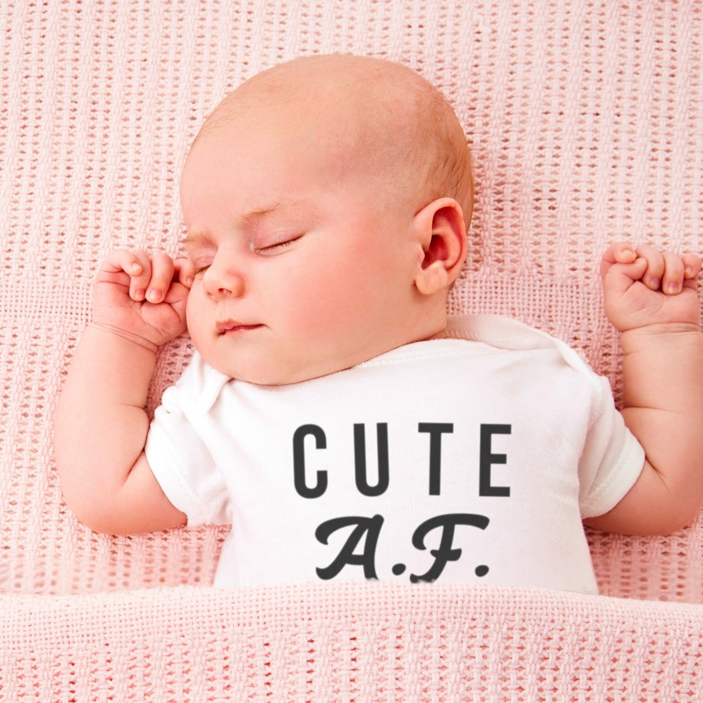 Baby wearing a onesie with 'CUTE A.F.' text on a pink blanket. A funny humorous irreverent baby outfit perfect for memorable baby photos or as a hilarious baby shower gift from Born Brash for cool parents.