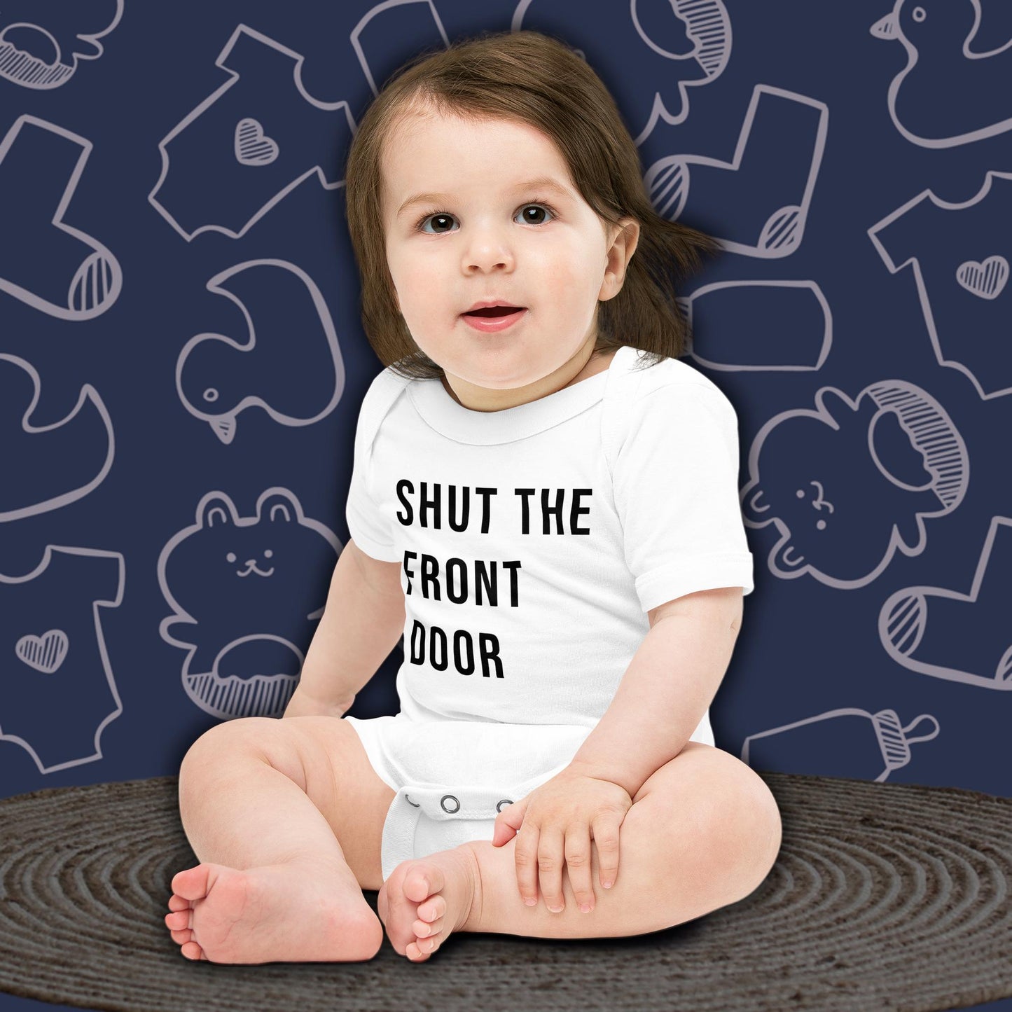 A cute baby with dark hair, sitting and looking forward, wearing a white Born Brash bodysuit that says 'Shut the Front Door'. The background is dark blue with white doodle patterns of baby bodysuits, baby toys, and baby bottles. Perfect for a funny newborn gift or pregnancy announcement.