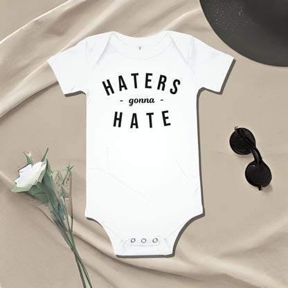 White baby bodysuit with "HATERS gonna HATE" text on draped fabric with accessories. A bold attitude baby clothes design that makes a sarcastic statement from Born Brash.