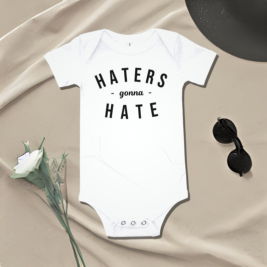 White baby bodysuit with "HATERS gonna HATE" text on draped fabric with accessories. A bold attitude baby clothes design that makes a sarcastic statement from Born Brash.