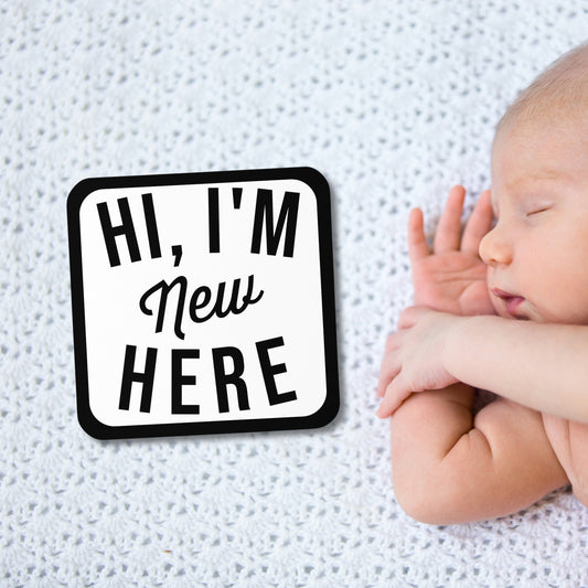 "HI, I'M New HERE" square photo prop sign placed next to a sleeping baby on a white blanket. A funny humorous newborn photo accessory perfect for capturing new baby photos or as a unique baby shower gift from Born Brash.