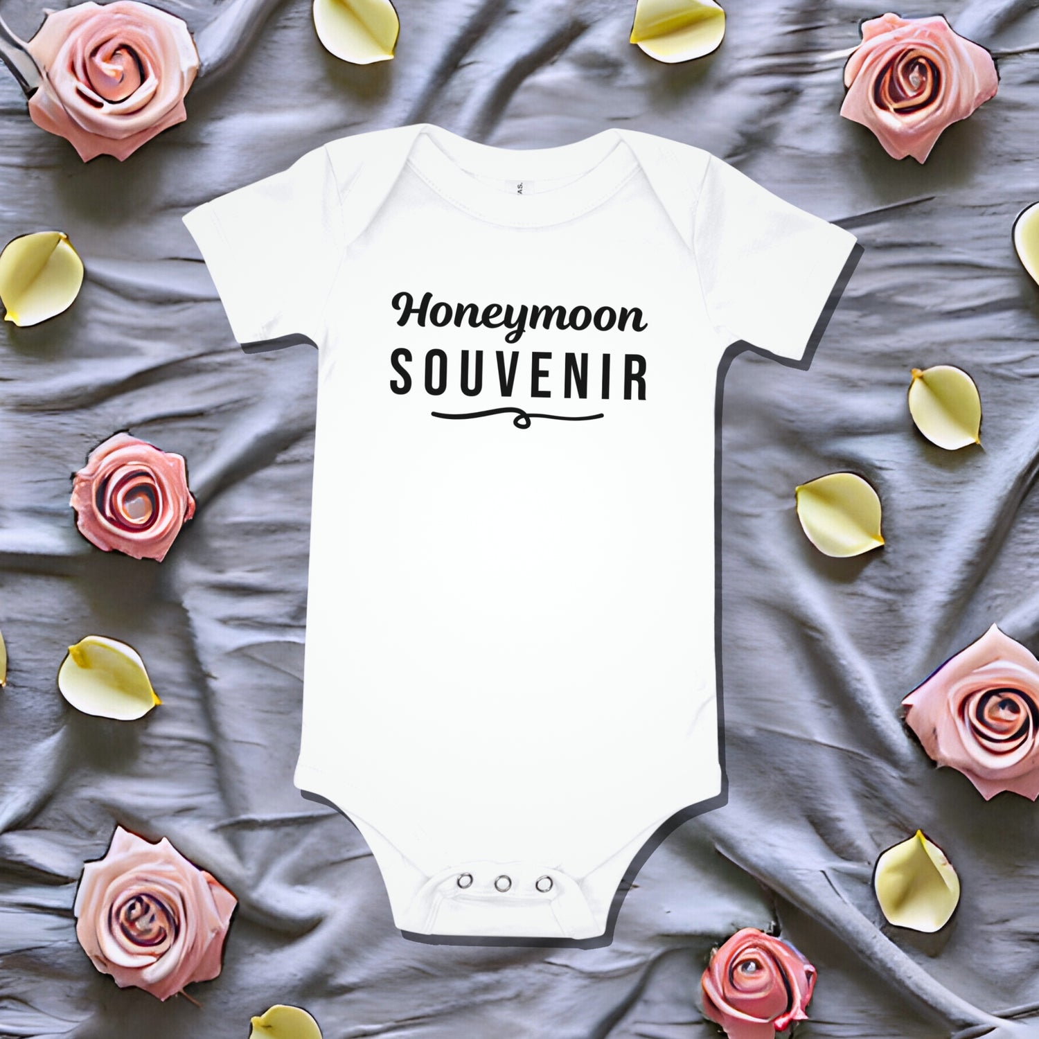 White baby bodysuit that says 'Honeymoon Souvenir' in black script, laid on a gray fabric background with pink rose petals. Funny newborn gift, unique baby shower idea, pregnancy announcement bodysuit.