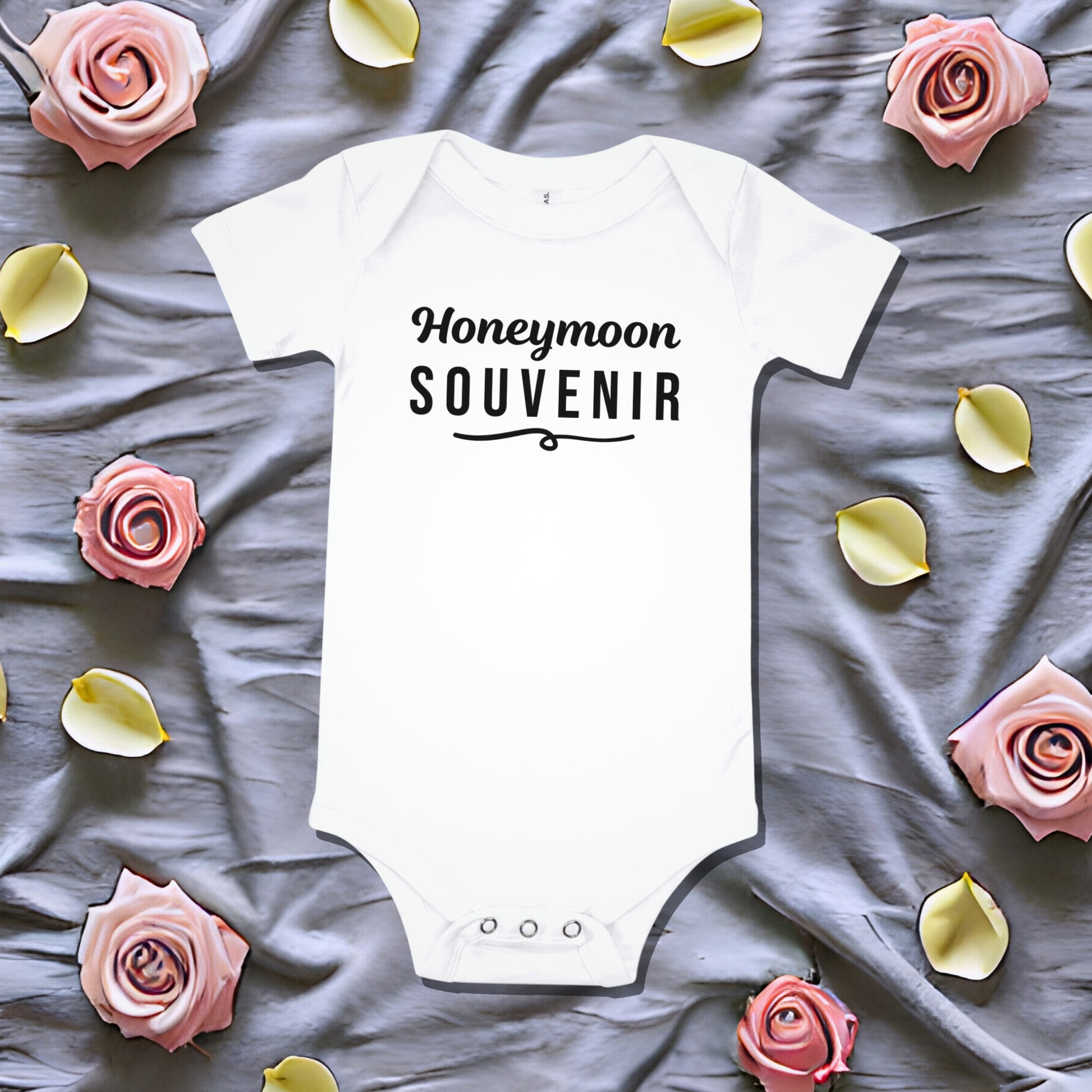 White baby bodysuit that says 'Honeymoon Souvenir' in black script, laid on a gray fabric background with pink rose petals. Funny newborn gift, unique baby shower idea, pregnancy announcement bodysuit.