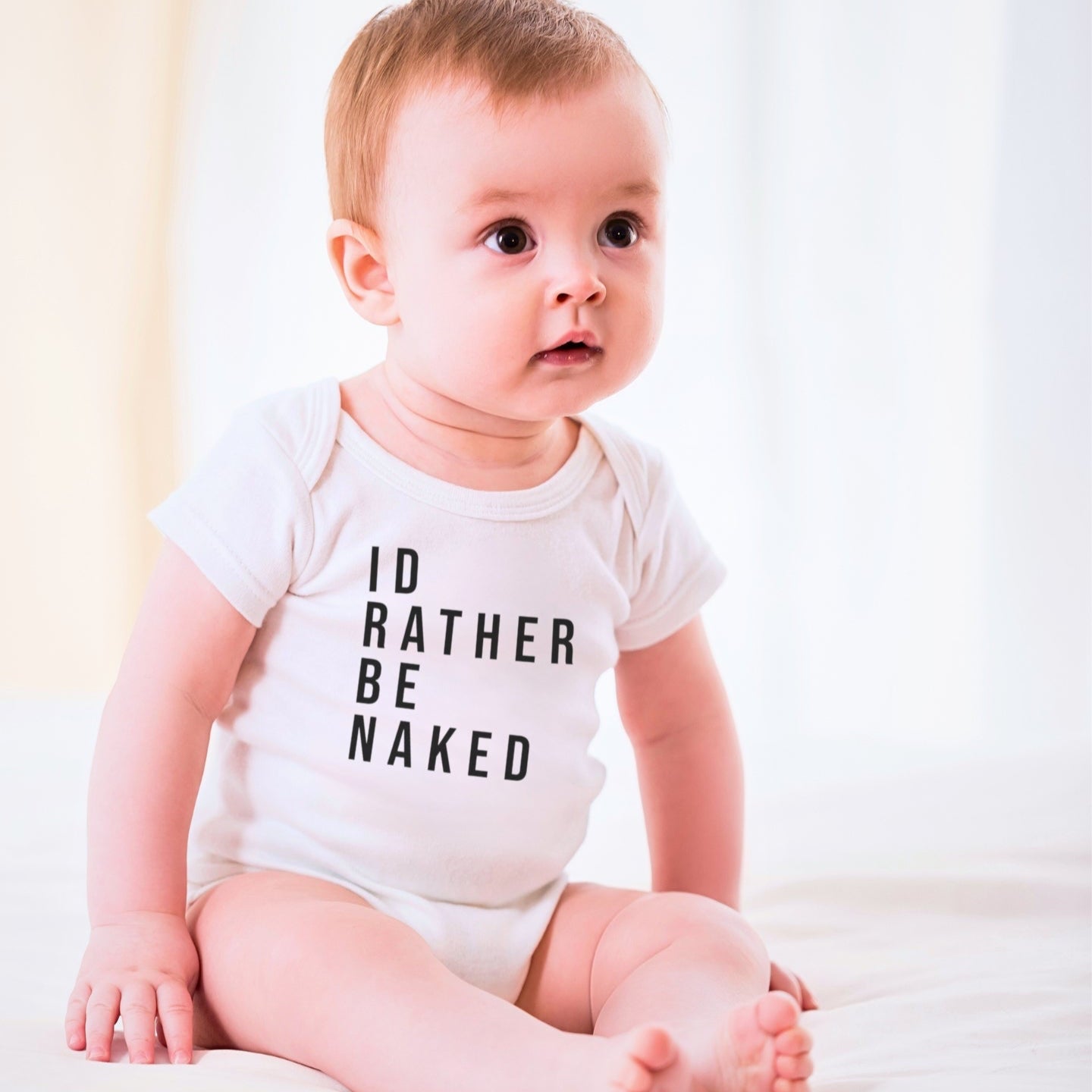 Baby wearing a white onesie with 'I D RATHER BE NAKED' text on a light background. A funny humorous irreverent baby outfit perfect for memorable baby photos or as a hilarious baby shower gift from Born Brash for cool parents.
