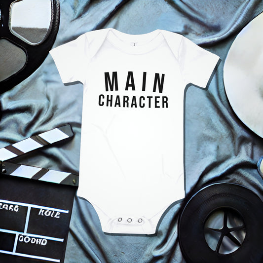 White baby bodysuit with "MAIN CHARACTER" text on a grey draped background with film production illustrations. A bold statement attitude baby clothes design for the star of the show from Born Brash.
