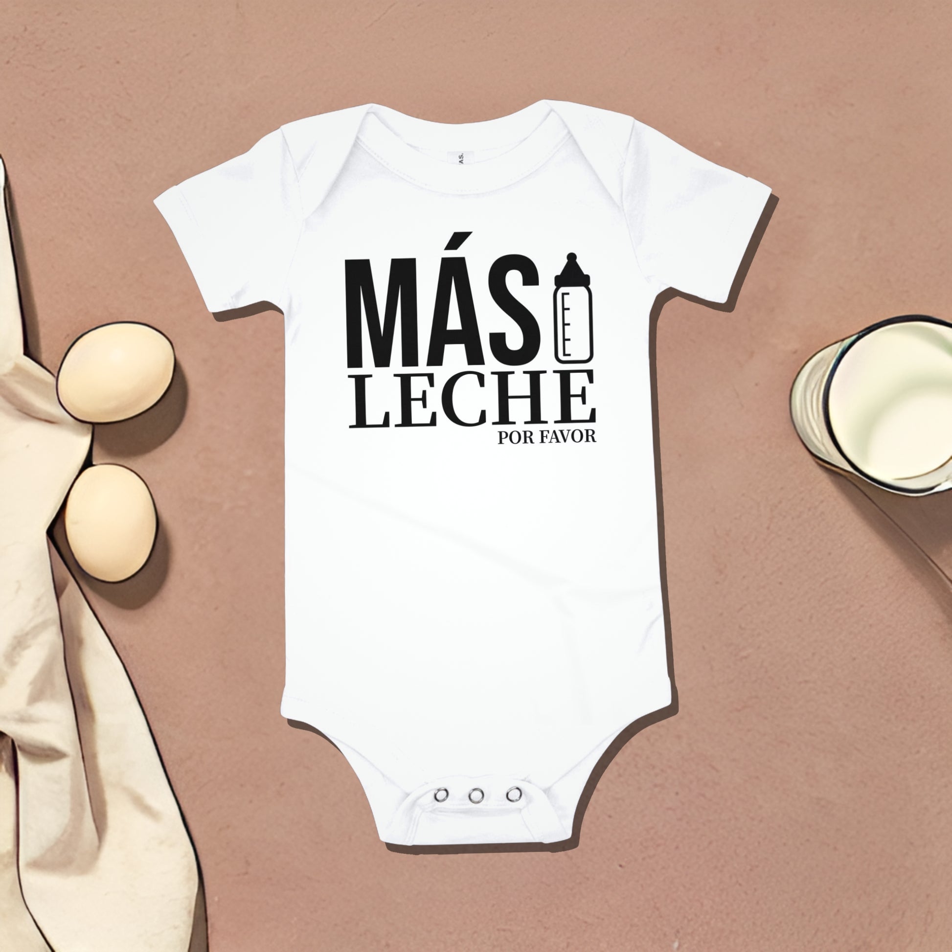 White baby bodysuit with "MÁS LECHE POR FAVOR" text and baby bottle graphic, on a dark brown background with egg and cup illustrations. A funny humorous baby clothes design about wanting more milk, perfect as a relatable baby shower gift from Born Brash.