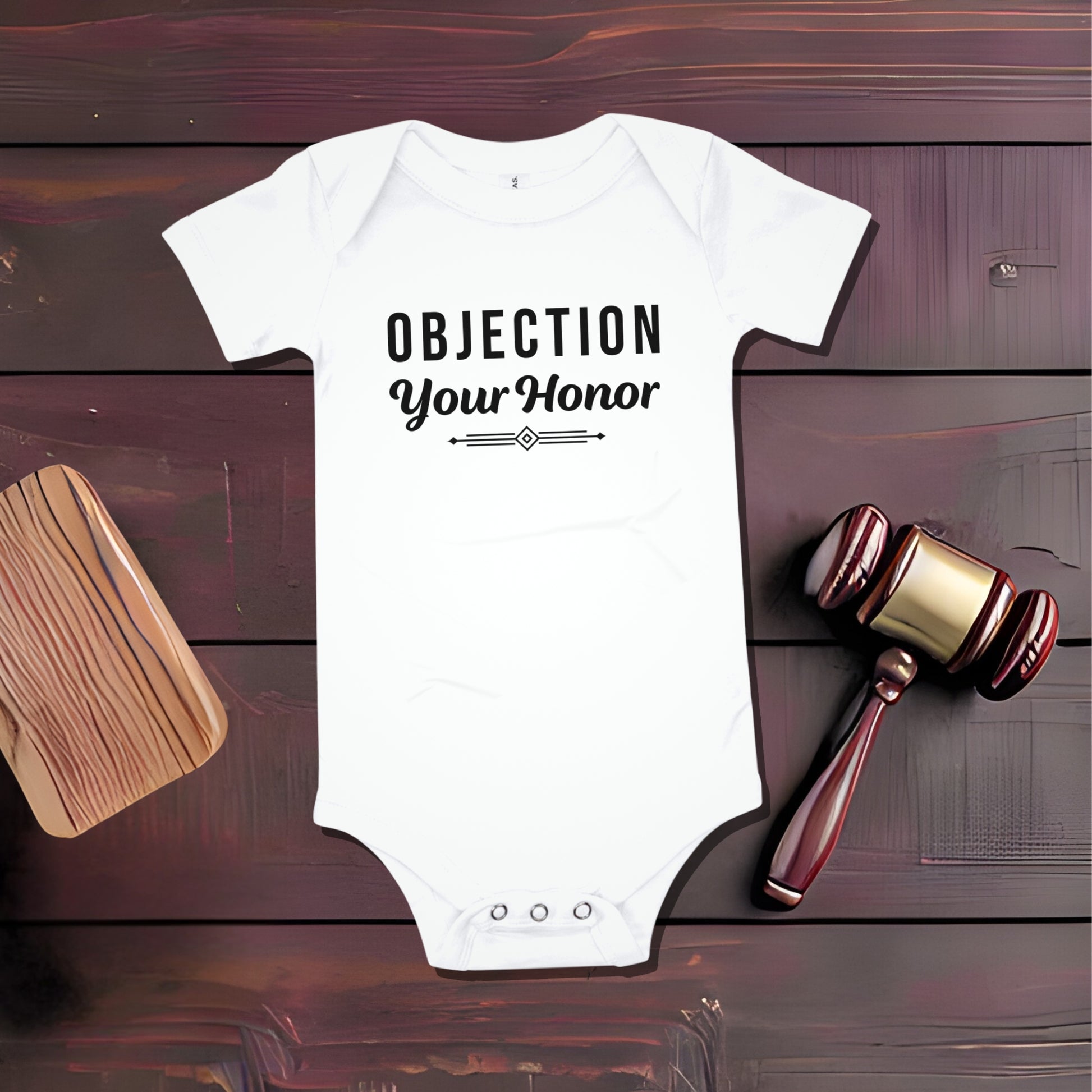 White baby bodysuit that says 'OBJECTION Your Honor' in black text, next to a wooden gavel and a sound block on a dark wood surface. Funny lawyer baby outfit, unique baby gifts, court themed baby clothes.