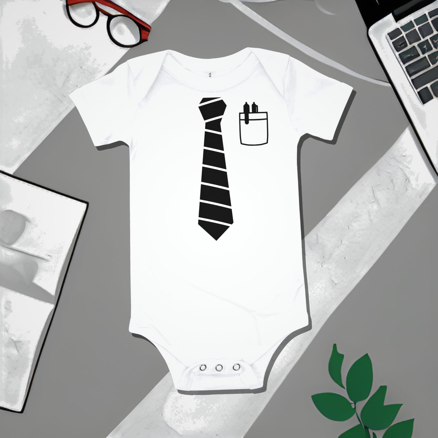 White baby bodysuit with a black striped tie graphic and shirt pocket with pens, on a gray surface with work illustrations. A funny humorous baby clothes design referencing a project manager, perfect as a unique baby shower gift from Born Brash.