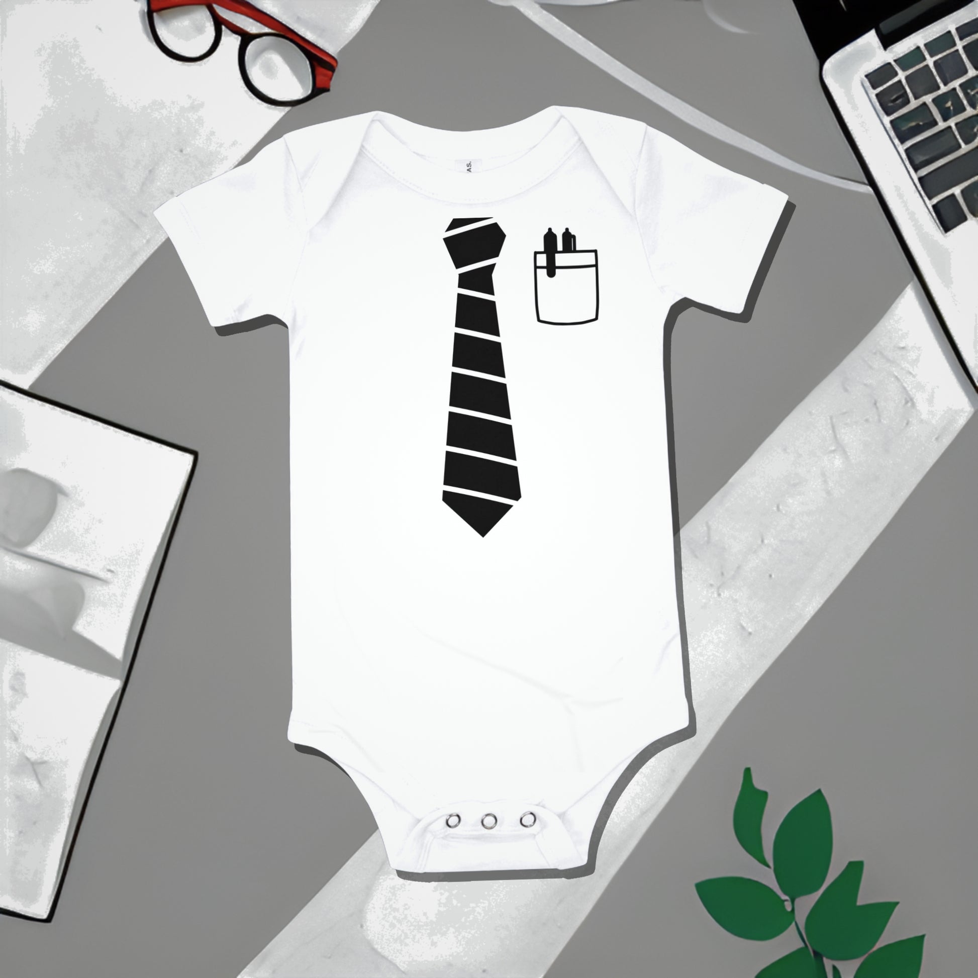 White baby bodysuit with a black striped tie graphic and shirt pocket with pens, on a gray surface with work illustrations. A funny humorous baby clothes design referencing a project manager, perfect as a unique baby shower gift from Born Brash.
