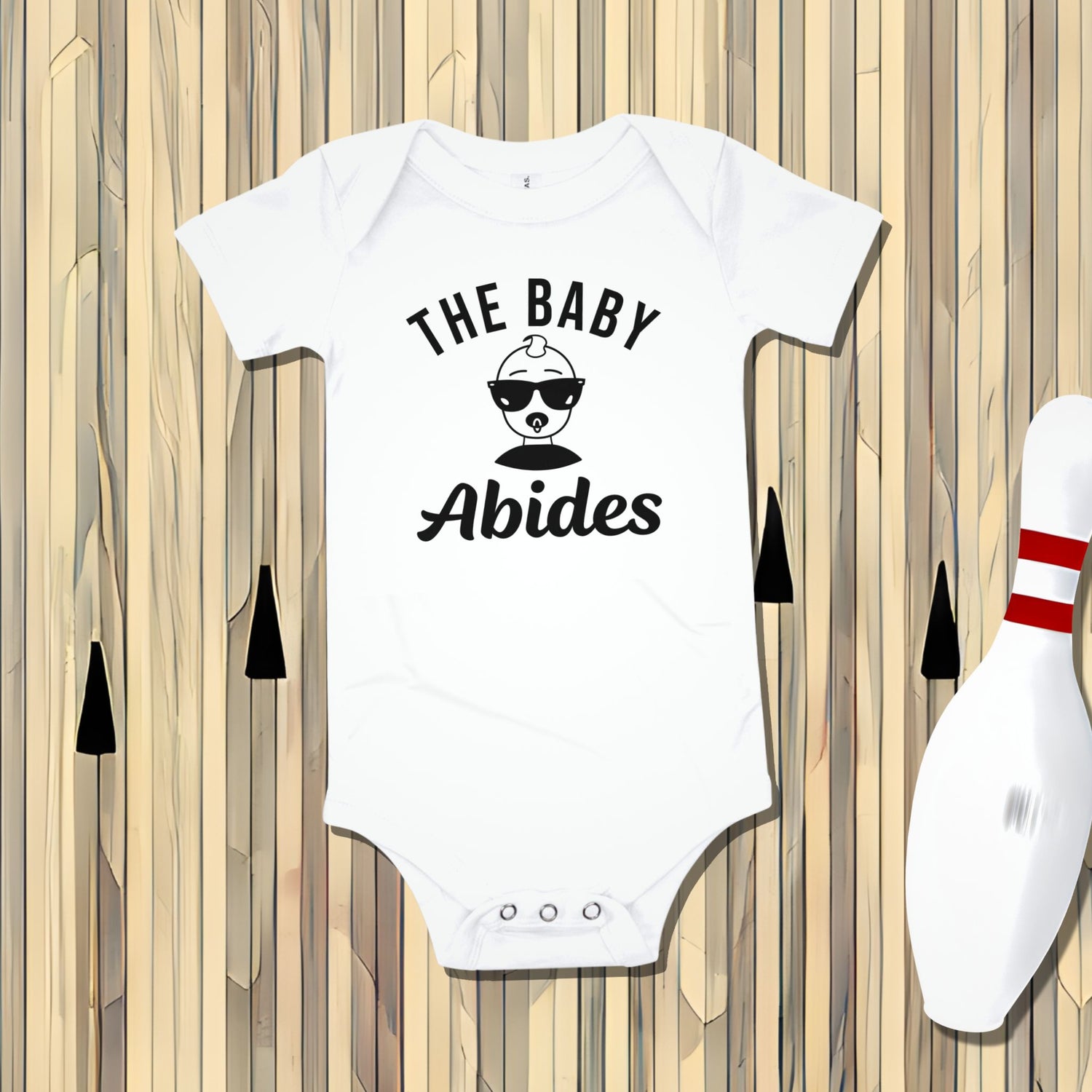 White Born Brash baby bodysuit that says 'THE BABY Abides' with a cool baby wearing sunglasses. The bodysuit is laid flat on a bowling alley lane background with a bowling pin. Funny baby clothes, The Big Lebowski inspired outfit, unique baby gift for fans.