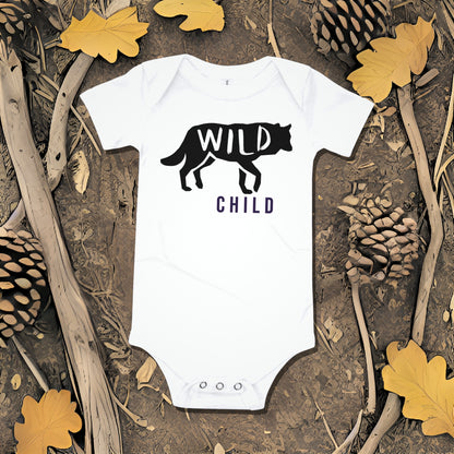 White baby bodysuit with a wolf silhouette graphic and "WILD CHILD" text on a background of bark, pinecones, and oak leaves. A bold unique baby clothes design perfect as a baby shower gift from Born Brash for little wild ones.