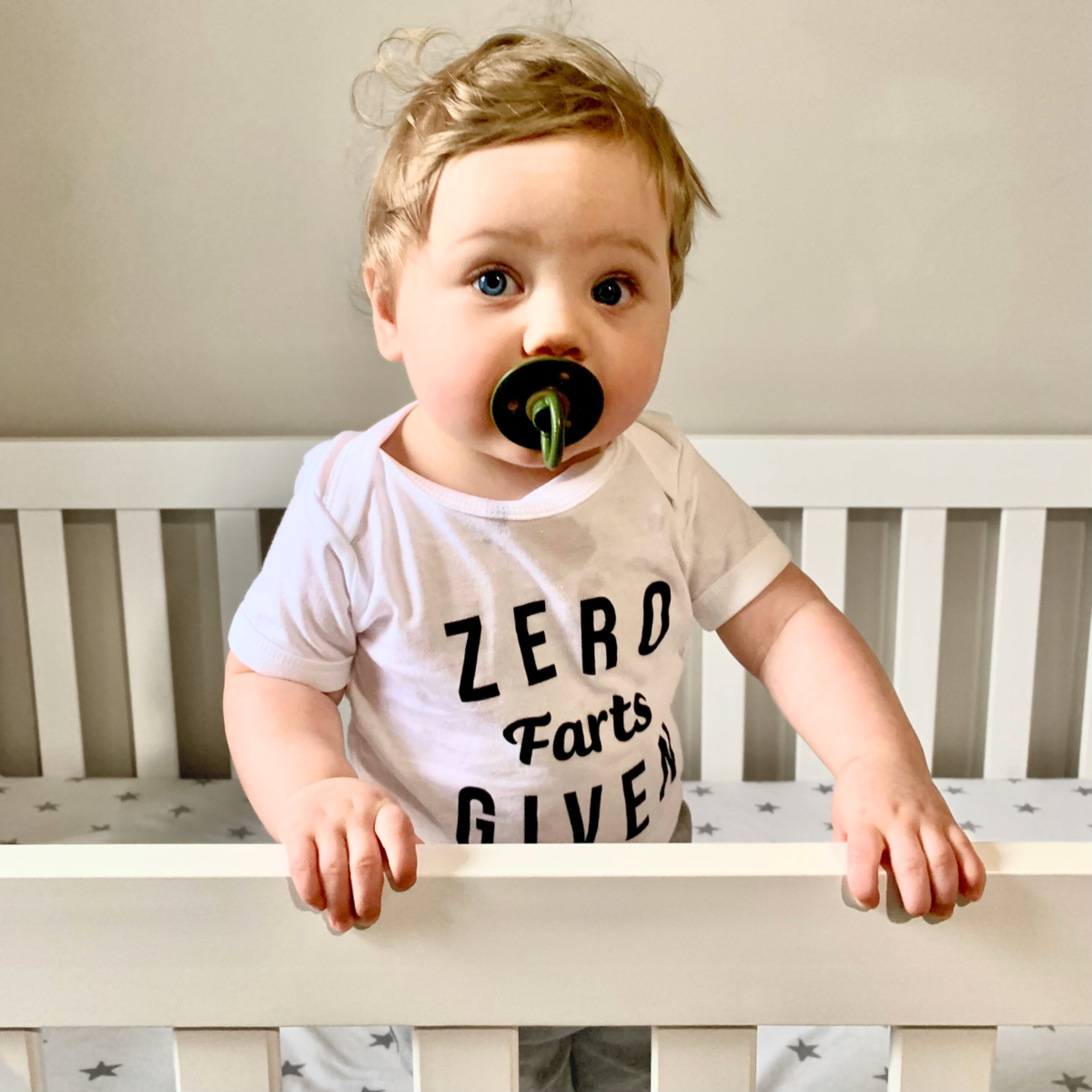 Baby wearing a white "ZERO Farts GIVEN" bodysuit and holding onto a crib rail. A funny humorous irreverent baby outfit perfect for memorable baby photos or as a hilarious baby shower gift from Born Brash for cool parents.