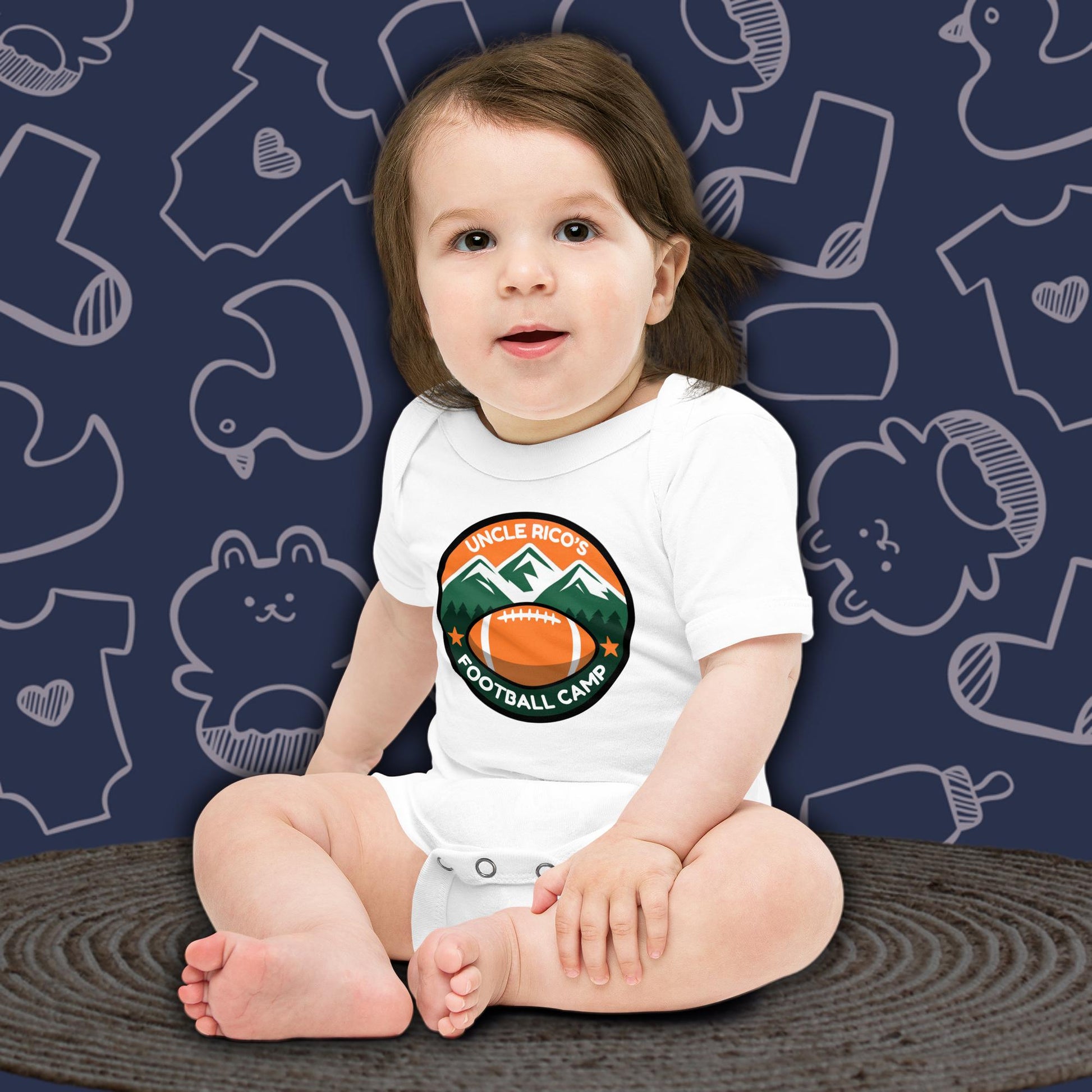 A cute baby with dark hair, sitting and looking forward, wearing a white Born Brash bodysuit that says 'Uncle Rico's Football Camp' with an illustration of a football and mountains. The background is dark blue with white doodle patterns of baby bodysuits, baby toys, and baby bottles. Funny football themed baby clothes. Perfect baby gift for football fans or Napoleon Dynamite fans.