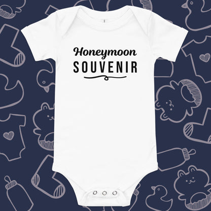 Funny, white baby bodysuit featuring the playful text "Honeymoon Souvenir" underlined against a whimsical, blue backdrop of baby toys, bodysuits, and bottles.