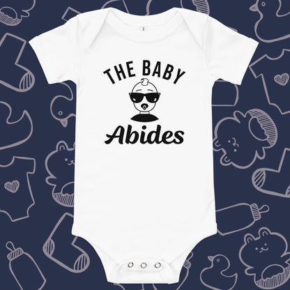 Funny, white baby bodysuit featuring the playful text "The Baby Abides" and an illustration of a cool baby wearing sunglasses against a whimsical, blue backdrop of baby toys, bodysuits, and bottles.